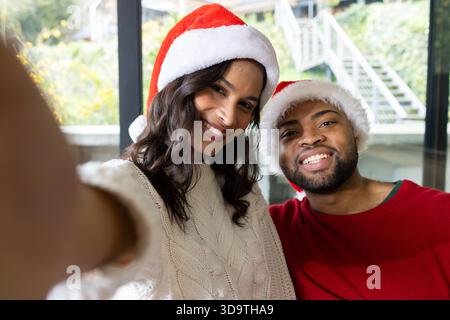 Diverse Paare mit Weihnachtsmützen und Strickpullover, die Selfie im Wohnzimmer mit Smartphone machen Stockfoto