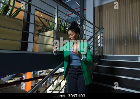 Afroamerikanerin, die auf einer Treppe steht und ihr Telefon in grünem Blazer mit Blick auf das Büro hält Stockfoto
