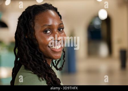 Afroamerikanische Frau mit grünem Oberteil, die sich der Kamera zuwendet und in der Lobby mit Hängeleuchten lächelt Stockfoto