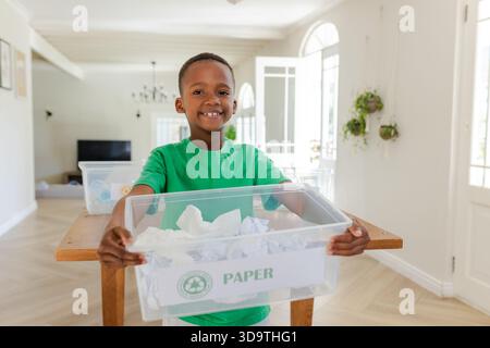 Afroamerikanisches männliches Kind trägt ein grünes T-Shirt mit klarem PAPIERKORB und BESCHRIFTETEM PAPIER zu Hause Stockfoto
