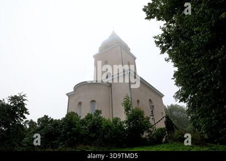 Beige Steinkirche mit Kuppelturm und Kreuz über Laubbäumen im nebeligen Morgenlicht auf der Insel Suomenlinna, Helsinki, Finnland, eine flache Aussicht. Stockfoto