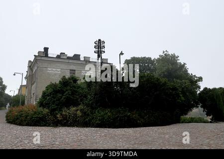Historisches Gipsgebäude und Metallgitterturm mit runden Rahmen über dichten Sträuchern an einem nebeligen Morgen auf der Insel Suomenlinna, Helsinki, Finnland. Stockfoto