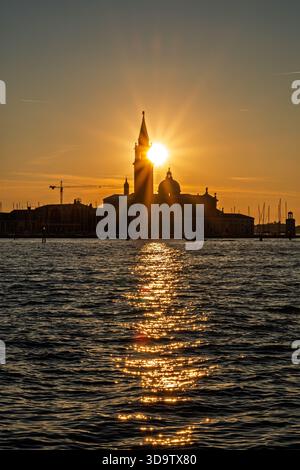 Vertikale Komposition des Sonnenuntergangs von Venedig mit strahlender Sonne, die über dem Kirchturm von San Giorgio Maggiore ausgerichtet ist, und goldener Lichtpfad, der sich über dunkle Wellen erstreckt. Stockfoto