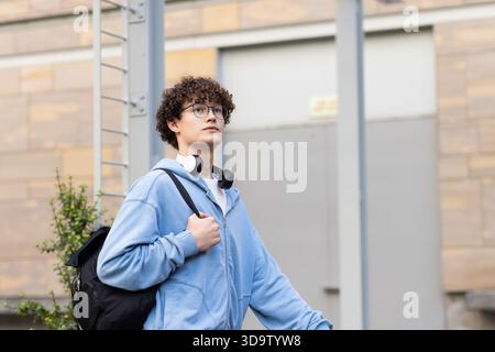 Mann, der neben der Steinfassade steht, trägt einen hellblauen Hoodie mit Brille, Kopfhörern und Rucksack Stockfoto