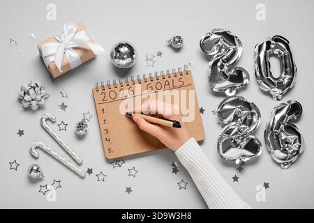 Frau mit Figur 2026, Geschenkbox und Weihnachtsdekor, die To-do-Liste auf grauem Hintergrund schreibt Stockfoto