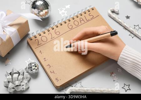 Frau mit Geschenkbox und Weihnachtsdekor, die 2026 To-do-Liste auf grauem Hintergrund schreibt Stockfoto