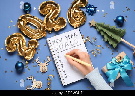 Frau mit Figur 2026, Geschenkbox und Weihnachtsdekor, die To-do-Liste auf blauem Hintergrund schreibt Stockfoto