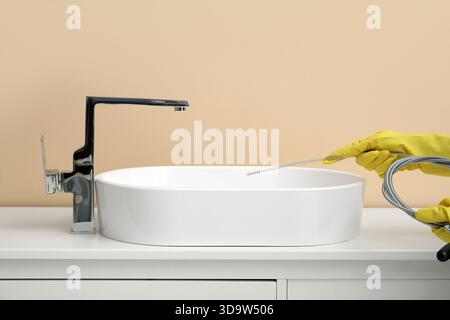 Frau in Gummihandschuhen, die verstopften Abfluss mit Klempnerschlange im Badezimmer reinigen Stockfoto