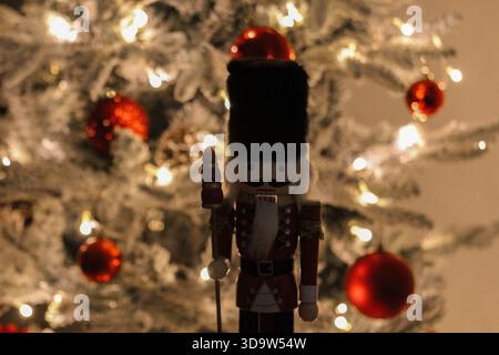 Ein klassischer hölzerner Nussknacker steht vor einem sanft beleuchteten Weihnachtsbaum, der mit roten Ornamenten und mattierten Zweigen verziert ist und ein warmes, festliches atmos schafft Stockfoto