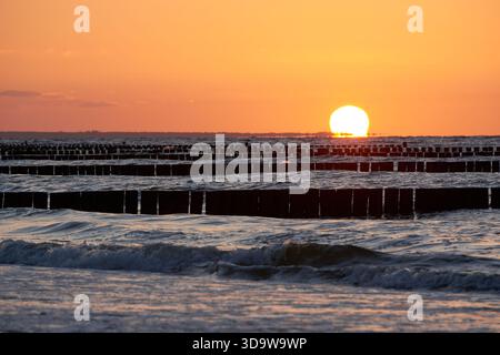 Die Sonne untergeht über der Ostsee und reflektiert warmes orangefarbenes Licht auf den Wellen, während Reihen von hölzernen Wellenbrechern rhythmische Linien im Wasser und al erzeugen Stockfoto