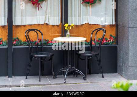 Melbourne, VIC, Australien. Dezember 2025. - Ein einladender Kaffeetisch im Freien mit zwei schwarzen Stühlen und einer Vase mit gelben Blumen sitzt auf einem Bürgersteig Stockfoto