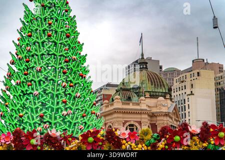 Melbourne, VIC, Australien. Dezember 2025. - Ein großer, hell erleuchteter Weihnachtsbaum und farbenfrohe Blumendekorationen zieren eine Stadtstraße mit einem Histo Stockfoto
