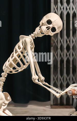 Das Skelett lehnt sich an ein Industrietor. Symbolisiert dunkle Wissenschaft, psychische Gesundheit, Burnout und existenzielle Ironie. Stockfoto