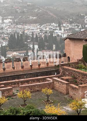 Regnerische Aussicht von den Alhambra-Gärten in Granada, Spanien, mit historischen Festungsmauern, Herbstbäumen und nebeliger Albaicin-Stadtlandschaft. Stockfoto