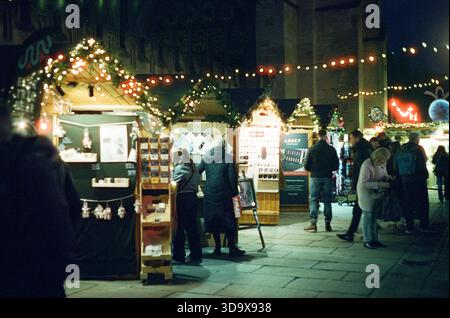 Bath, England - 3. Dezember 2025: Bath Christmas Market, mit Verkaufsständen, Verkaufsständen und Käufern. Aufgenommen nach Einbruch der Dunkelheit auf Spezialfilm. Stockfoto