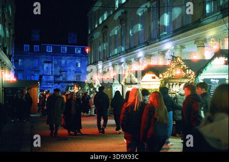 Bath, England - 3. Dezember 2025: Bath Christmas Market, mit Verkaufsständen, Verkaufsständen und Käufern. Aufgenommen nach Einbruch der Dunkelheit auf Spezialfilm. Stockfoto