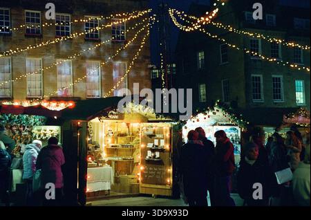 Bath, England - 3. Dezember 2025: Bath Christmas Market, mit Verkaufsständen, Verkaufsständen und Käufern. Aufgenommen nach Einbruch der Dunkelheit auf Spezialfilm. Stockfoto