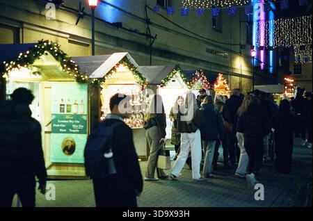 Bath, England - 3. Dezember 2025: Bath Christmas Market, mit Verkaufsständen, Verkaufsständen und Käufern. Aufgenommen nach Einbruch der Dunkelheit auf Spezialfilm. Stockfoto