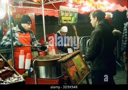 Bath, England - 3. Dezember 2025: Bath Christmas Market, mit Verkaufsständen, Verkaufsständen und Käufern. Aufgenommen nach Einbruch der Dunkelheit auf Spezialfilm. Stockfoto