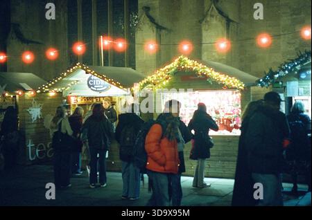 Bath, England - 3. Dezember 2025: Bath Christmas Market, mit Verkaufsständen, Verkaufsständen und Käufern. Aufgenommen nach Einbruch der Dunkelheit auf Spezialfilm. Stockfoto