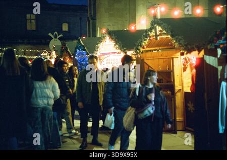 Bath, England - 3. Dezember 2025: Bath Christmas Market, mit Verkaufsständen, Verkaufsständen und Käufern. Aufgenommen nach Einbruch der Dunkelheit auf Spezialfilm. Stockfoto