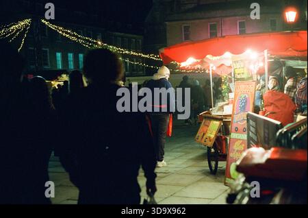 Bath, England - 3. Dezember 2025: Bath Christmas Market, mit Verkaufsständen, Verkaufsständen und Käufern. Aufgenommen nach Einbruch der Dunkelheit auf Spezialfilm. Stockfoto