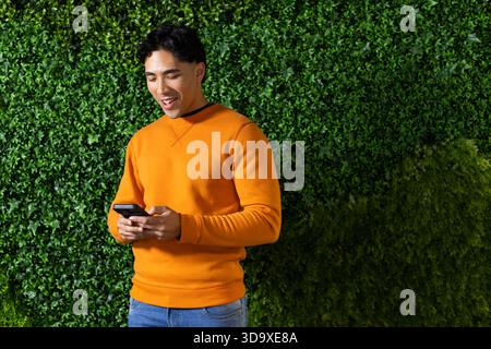 Asiatischer Mann, der an der Hecke im Garten steht und lächelt, während er ein Smartphone mit orangefarbenem Sweatshirt hält Stockfoto