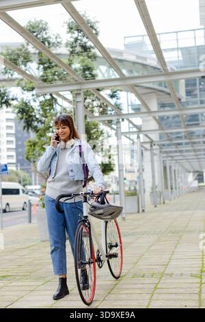 Frau, die mit dem Fahrrad unter einem Glasdach entlang des Bürgersteigs unterwegs ist, während sie Smartphone und Helm hält Stockfoto