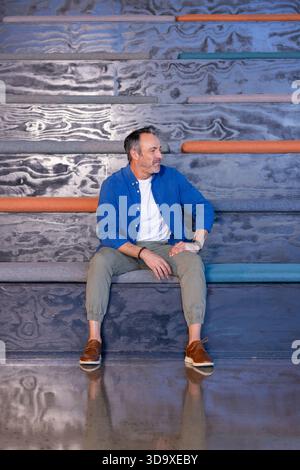 Reifer Mann sitzt auf gestuften Bänken mit farbigen Kissen und Armbanduhr über Betonboden Stockfoto
