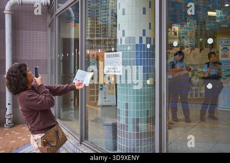 Hongkong, China. Dezember 2025. Eine Wählerin fotografiert ihre Wahlurkunde vor einem Wahllokal und Polizeibeamte überwachen den reibungslosen Ablauf der Wahlen in Tin Shui Wai. Die Parlamentswahlen 2025 in Hongkong finden am 7. Dezember 2025 statt, um die 90 Mitglieder des 8. Hongkonger Legislativrates zu wählen. Quelle: SOPA Images Limited/Alamy Live News Stockfoto