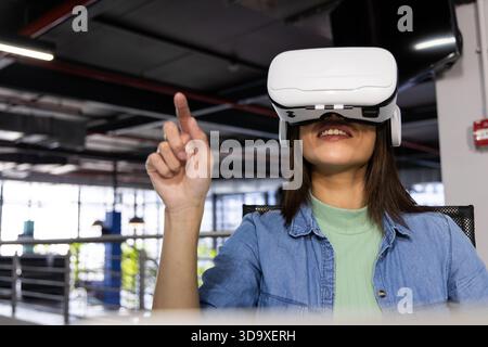 Frau sitzt mit weißem VR-Headset und interagiert mit virtuellen Steuerungen im Großraumbüro Stockfoto