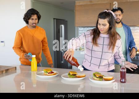 Verschiedene Freunde gießen Ketchup auf Burger auf der Insel-Arbeitsplatte in der Küche mit blauem Reinigungstuch Stockfoto