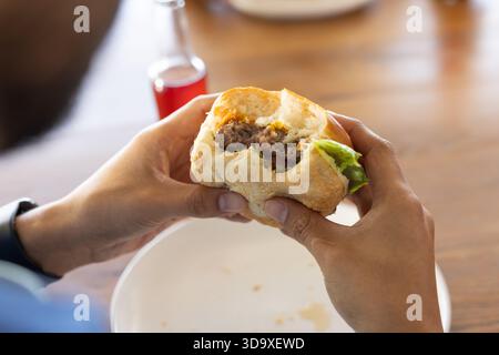 Mittelalter Mann, der halbgefressenen Burger über Teller auf Holztisch hält, rote Flasche, Armbanduhr zeigt Stockfoto