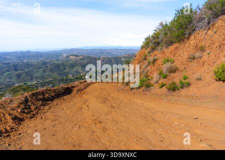 Die sich windende rötliche Schotterstraße führt durch trockenes Brushland und zeigt weitläufige grüne Hügel und ferne Berge unter einem hellblauen Himmel. Stockfoto