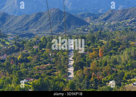 Blick von oben auf eine ruhige Straße, die sich durch ein lebendiges, baumgesäumtes Wohntal mit majestätischen Bergen in der Ferne schlängelt. Stockfoto