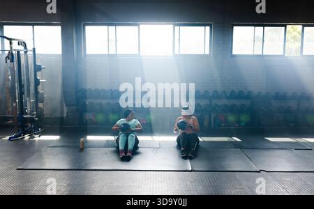 Verschiedene Freundinnen sitzen auf Matten und halten Medizinbälle in sportlicher Kleidung in der Nähe von Kurzhanteln Stockfoto