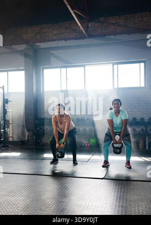 Verschiedene Freundinnen, die Trainingsausrüstung tragen, heben Wasserkettlebells auf Matten im Fitnessstudio bei Sonnenlicht an Stockfoto