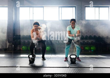 Vielseitiges weibliches Duo in sportlicher Kleidung, klatscht und reibt Kreide in die Hände von Kettlebells im Fitnessstudio Stockfoto