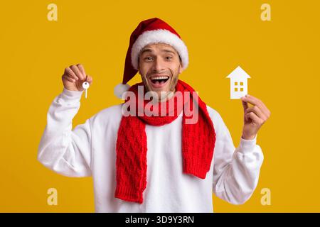 Aufgeregter Mann, der Weihnachten feiert und Keys und ein Hausmodell hält Stockfoto