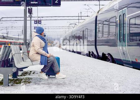 Reife Reiserin mit Koffer sitzt auf einer Bank an einem verschneiten Bahnhof und wartet auf die Ankunft des Zuges, in warmer Winterkleidung gekleidet und fängt ihn ein Stockfoto