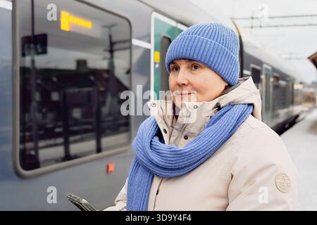 Eine Frau mittleren Alters, die in Winterkleidung unterwegs ist, steht am Bahnhof, hält Smartphone, wartet auf den Zug, mit städtischen Verkehrselementen im Stockfoto