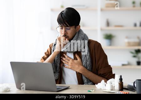 Mann mit Schal sitzt tagsüber an einem Schreibtisch und hustet, während er auf einen Laptop schaut Stockfoto