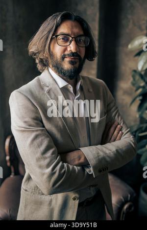 Porträt eines selbstbewussten Geschäftsmannes, der mit gefalteten Armen in einem stilvollen Büro steht und Führungsstärke, Professionalität und Vertrauen in einem Modus ausdrückt Stockfoto
