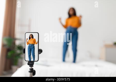 Frau tanzt fröhlich, während sie sich mit dem Smartphone auf einem Stativ in ihrem Schlafzimmer aufnimmt Stockfoto