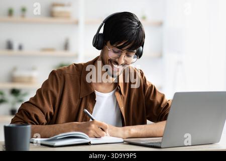 Mann, der zu Hause mit Kopfhörern studiert, während er Notizen in einem Notebook schreibt und einen Laptop in einem gut beleuchteten Raum benutzt Stockfoto