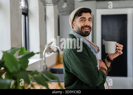Indischer Mann, der in der Küche steht und sich gegen die Theke lehnt, hält Becher neben Topfpflanzen, Kopierraum Stockfoto