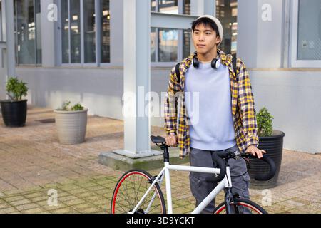 Asiatischer Mann, der neben weißem Fahrrad mit Kopfhörern auf dem Gehweg vor dem Gebäude steht, Kopierraum Stockfoto