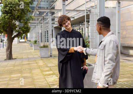 Verschiedene männliche Freunde schütteln sich die Hände auf dem Campus plaza in der Nähe von Glasdach, einer im Kleid mit Kappe Stockfoto