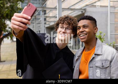 Verschiedene männliche Freunde im Kleid machen Selfie auf dem Campus mit Mortarboard und rotem Smartphone Stockfoto