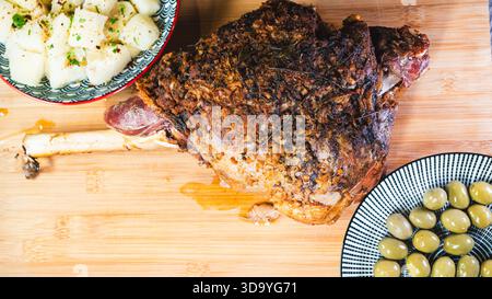 Gebratene Lammkeule auf einem Brett mit Beilagen: Oliven und Maniok-Pommes. Golden, gewürzt, perfekt für ein komplettes und herzhaftes festliches Essen. Stockfoto
